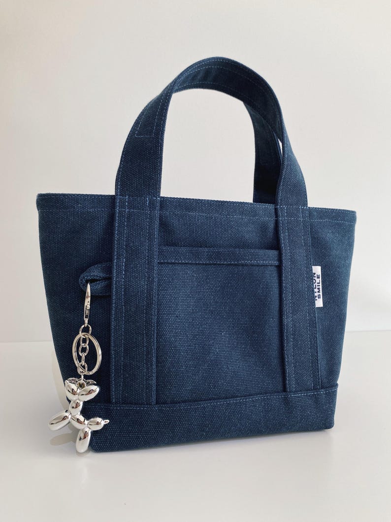 May include: A dark blue canvas tote bag with two top handles and a front slip pocket. A small white rectangular tag on the side reads NYLON SMILE. A silver-colored metallic balloon dog keychain is attached to the side of the bag by a metal ring.