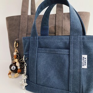 May include: A small dark blue canvas tote bag with two handles and a white rectangular label reading NYLON SMILE. A silver balloon dog keychain hangs from the side. Behind it sits a larger brown canvas tote bag with a beaded eight ball charm.