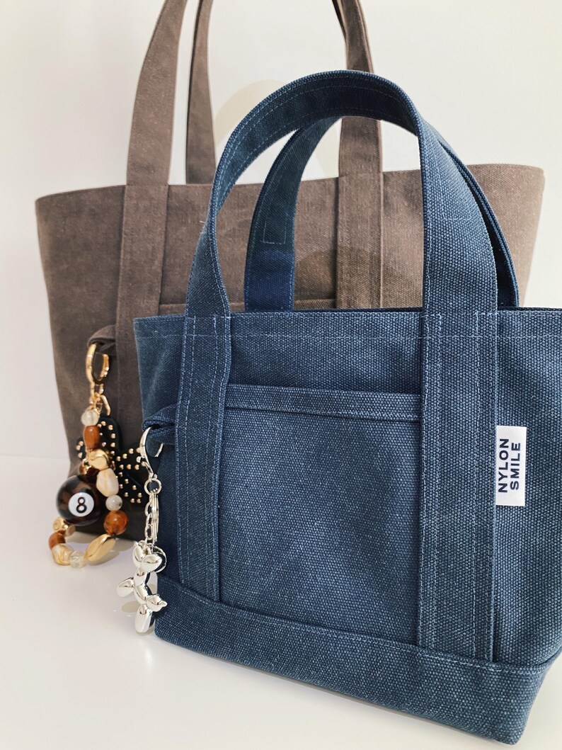 May include: A small dark blue canvas tote bag with two handles and a white rectangular label reading NYLON SMILE. A silver balloon dog keychain hangs from the side. Behind it sits a larger brown canvas tote bag with a beaded eight ball charm.