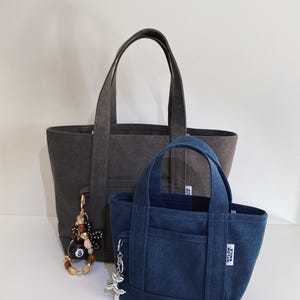 May include: A large charcoal gray tote bag and a smaller navy blue tote bag, both featuring dual handles, a front pocket, and a white rectangular tag with the text NYLON SMILE. The charcoal bag has a beaded charm, while the blue bag has a silver charm.