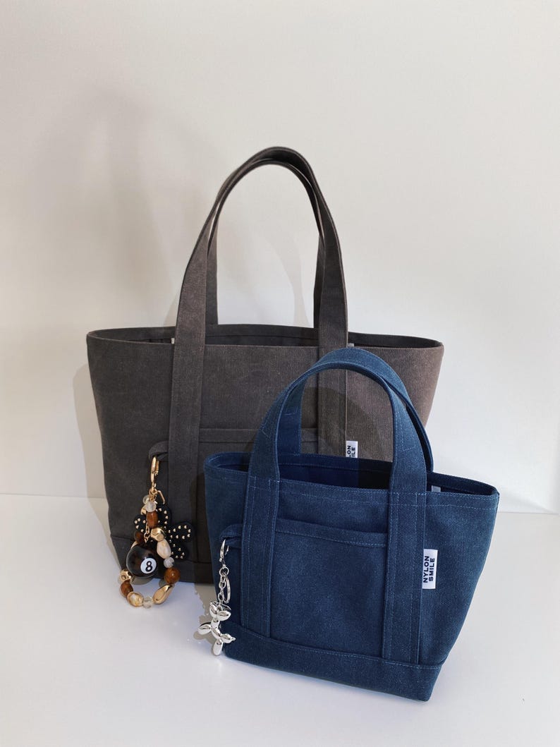 May include: A large charcoal gray tote bag and a smaller navy blue tote bag, both featuring dual handles, a front pocket, and a white rectangular tag with the text NYLON SMILE. The charcoal bag has a beaded charm, while the blue bag has a silver charm.