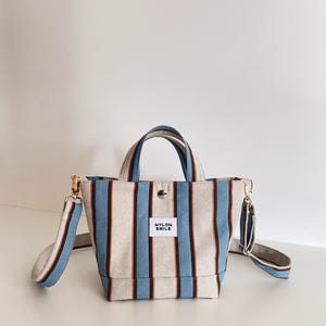 May include: A small tote bag featuring vertical stripes in light blue, dark brown, and off-white. The bag has two top handles, a detachable shoulder strap with gold-tone hardware, a snap button closure, and a white label reading NYLON SMILE.