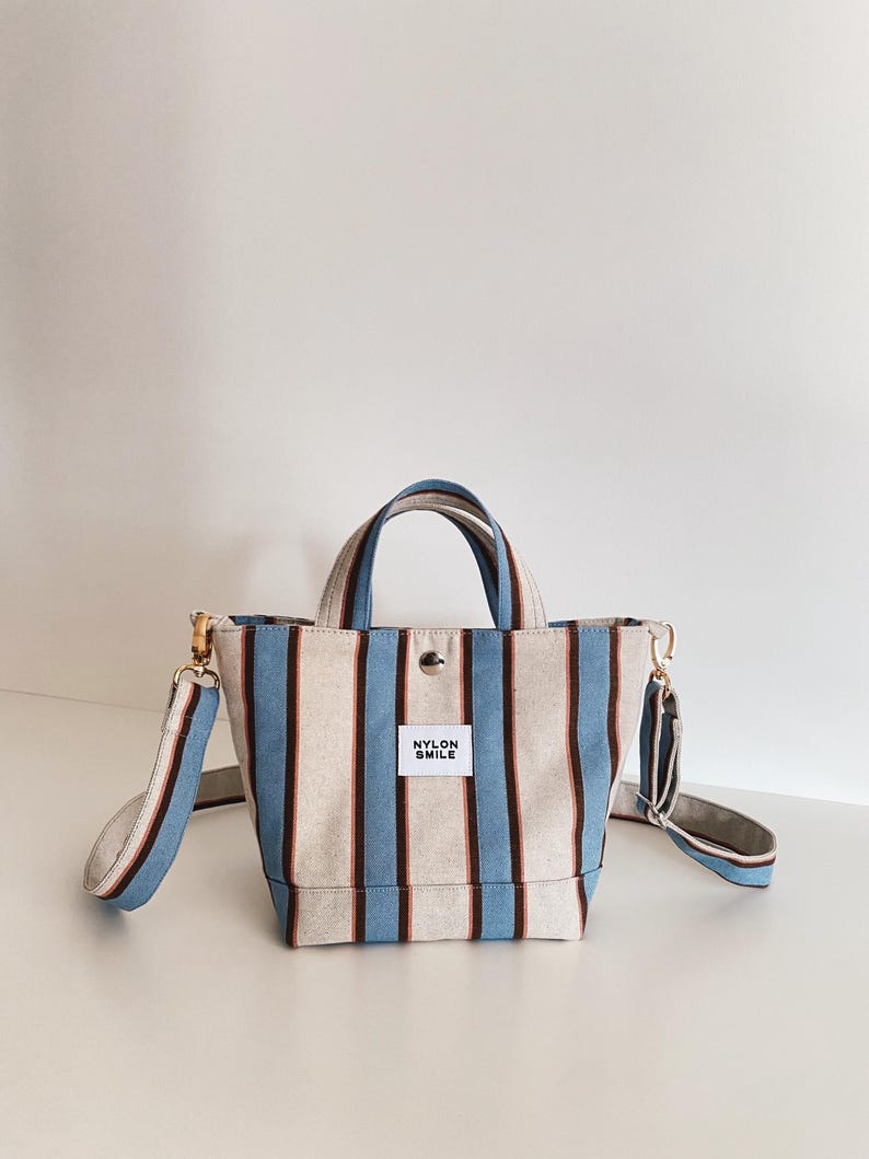 May include: A small tote bag featuring vertical stripes in light blue, dark brown, and off-white. The bag has two top handles, a detachable shoulder strap with gold-tone hardware, a snap button closure, and a white label reading NYLON SMILE.