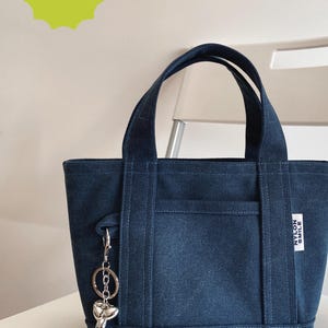 May include: A dark blue canvas tote bag with two top handles, an exterior front pocket, and a silver-toned balloon dog keychain attached to the side. A white rectangular label on the front reads NYLON SMILE. Text reads DIGITAL DOWNLOAD.