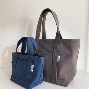 May include: A small blue canvas tote bag and a larger dark gray canvas tote bag, both featuring two top handles, an exterior front pocket, and a white rectangular fabric label with the black text NYLON SMILE stitched onto the front side.