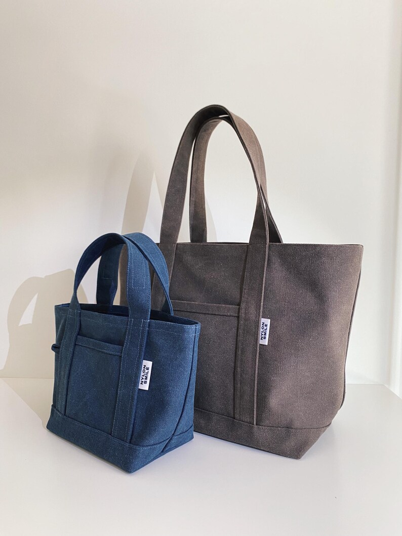 May include: A small blue canvas tote bag and a larger dark gray canvas tote bag, both featuring two top handles, an exterior front pocket, and a white rectangular fabric label with the black text NYLON SMILE stitched onto the front side.