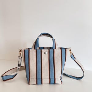 May include: A canvas tote bag featuring vertical stripes in light blue, dark brown, and off-white. The bag includes two top handles, a detachable shoulder strap with gold-toned metal hardware, and a single silver-toned snap button closure at the top.