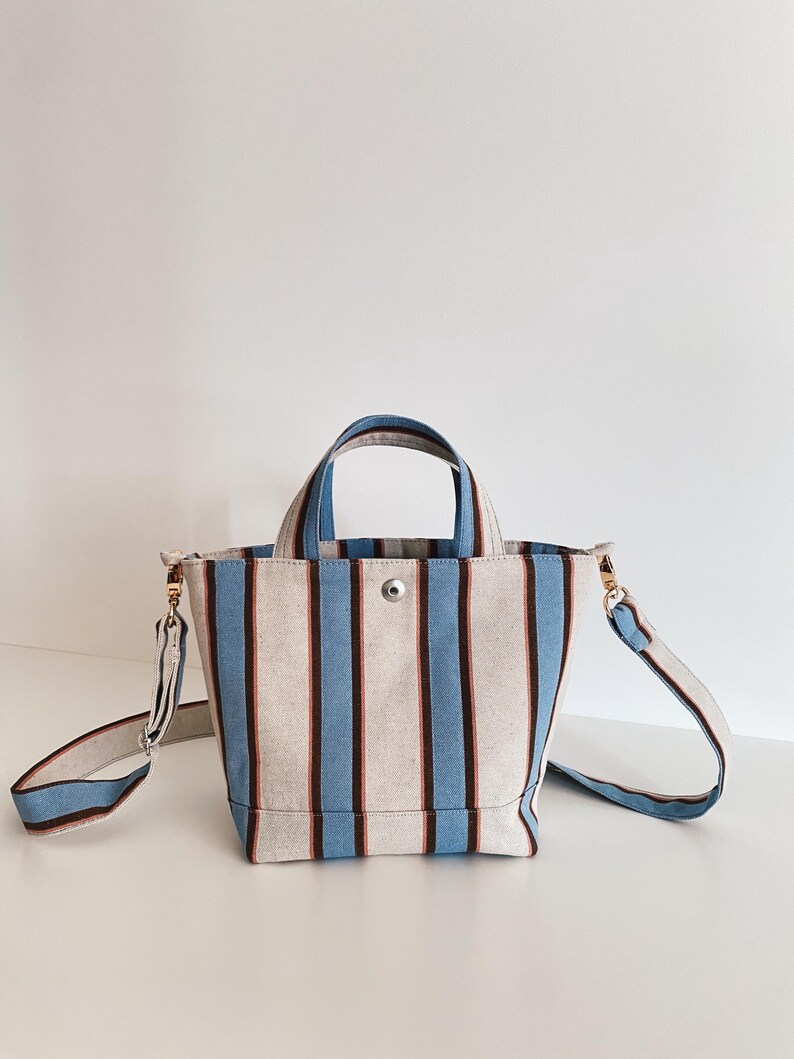 May include: A canvas tote bag featuring vertical stripes in light blue, dark brown, and off-white. The bag includes two top handles, a detachable shoulder strap with gold-toned metal hardware, and a single silver-toned snap button closure at the top.