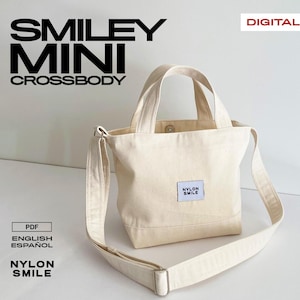 Peut inclure: Un sac bandoulière Smiley Mini de couleur crème avec une poignée supérieure et une bandoulière réglable. Le sac porte une petite étiquette carrée avec l'inscription "Nylon Smile". Le texte "Smiley Mini Crossbody" est imprimé en noir en haut de l'image.