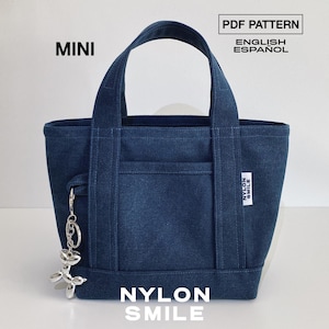 Op de afbeelding: Een donkerblauwe mini-tote bag met twee handvatten en een voorvak. Een zilveren ballonhond-sleutelhanger hangt aan de zijkant. Een wit label aan de zijkant bevat de tekst NYLON SMILE. De tekst bovenaan luidt MINI PDF PATTERN ENGLISH ESPAÑOL en NYLON SMILE.