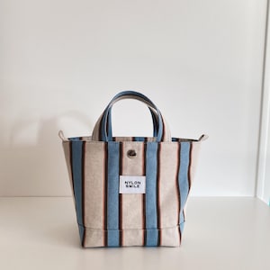May include: A small tote bag featuring vertical stripes in light blue, dark brown, and beige. The bag has two top handles, a silver-toned metal snap button closure, and a white rectangular fabric label on the front with the text NYLON SMILE.