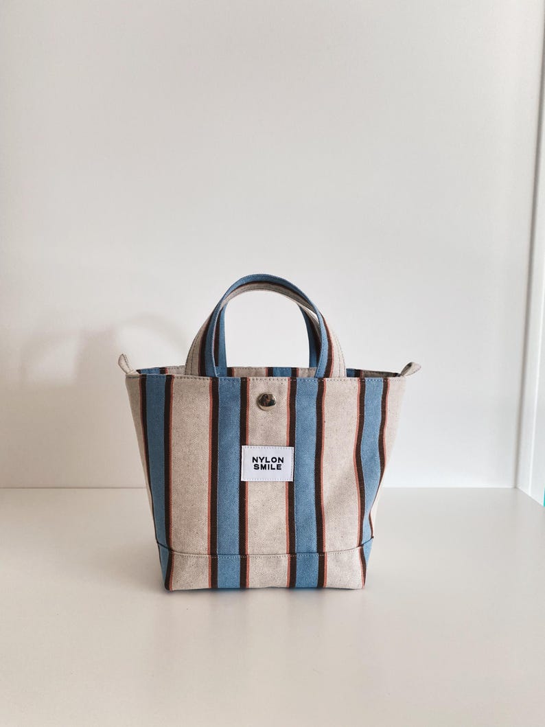 May include: A small tote bag featuring vertical stripes in light blue, dark brown, and beige. The bag has two top handles, a silver-toned metal snap button closure, and a white rectangular fabric label on the front with the text NYLON SMILE.