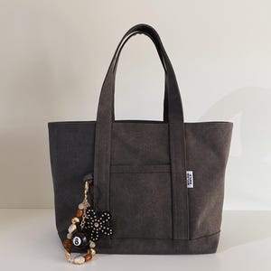 May include: A charcoal gray canvas tote bag with two top handles and a front pocket. A white label with the text NYLON SMILE is attached to the side. A decorative keychain featuring an eight ball, a black flower, and brown beads hangs from the bag.