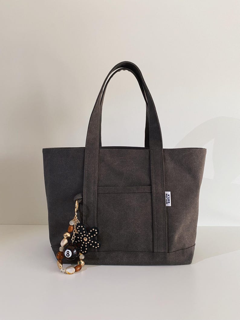 May include: A charcoal gray canvas tote bag with two top handles and a front pocket. A white label with the text NYLON SMILE is attached to the side. A decorative keychain featuring an eight ball, a black flower, and brown beads hangs from the bag.