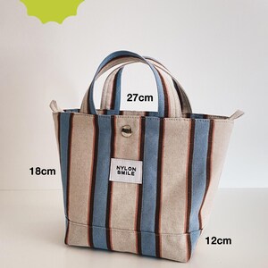 May include: A tote bag featuring vertical stripes in beige, light blue, and dark brown. The bag has two handles, a silver snap button, and a white label reading NYLON SMILE. Dimensions are 27cm wide, 18cm high, and 12cm deep. Text indicates skill level intermediate.