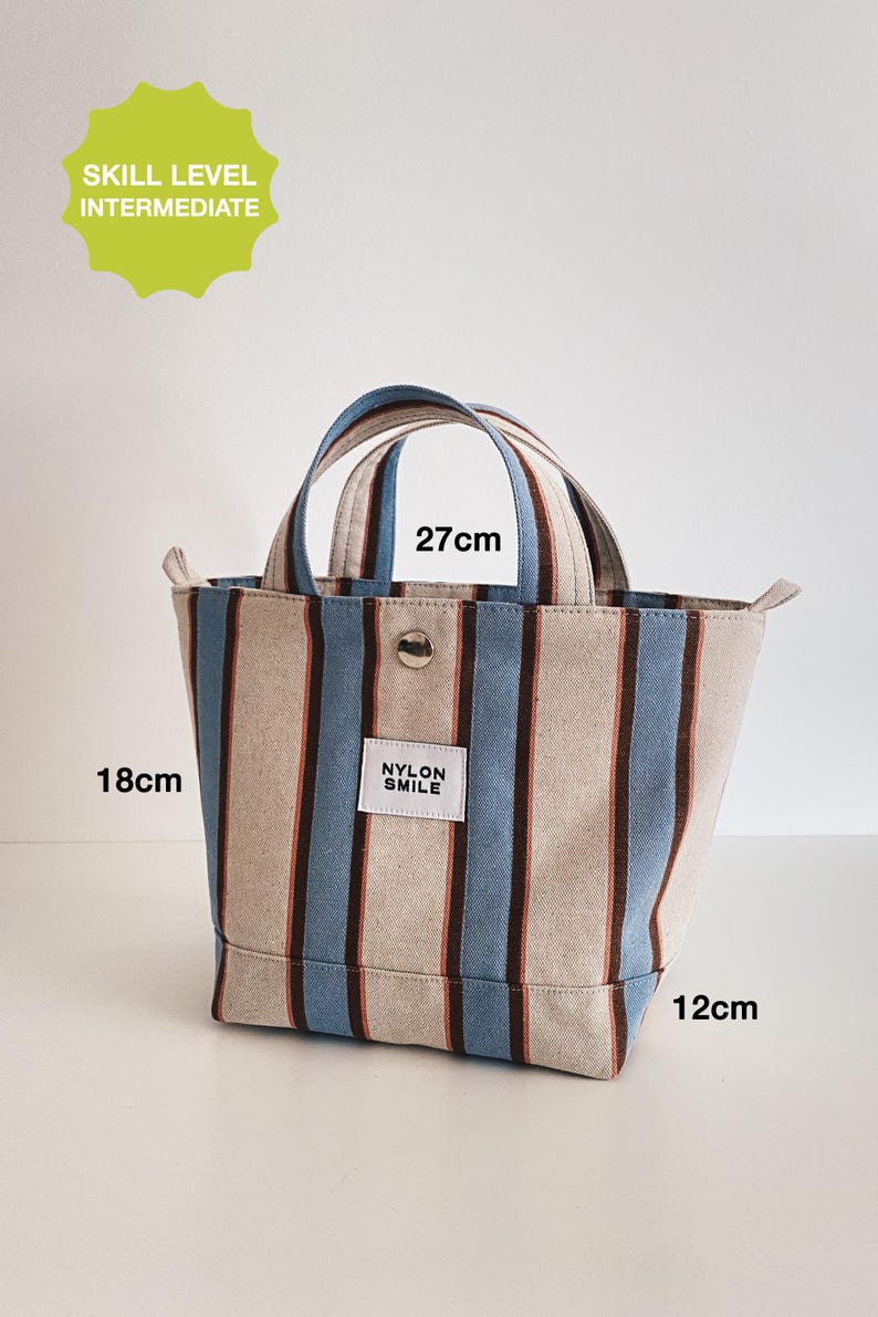 May include: A tote bag featuring vertical stripes in beige, light blue, and dark brown. The bag has two handles, a silver snap button, and a white label reading NYLON SMILE. Dimensions are 27cm wide, 18cm high, and 12cm deep. Text indicates skill level intermediate.