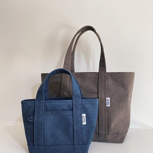 May include: A small blue denim tote bag and a larger brown canvas tote bag, both featuring two top handles, a front pocket, and a white rectangular label with the black text NYLON SMILE stitched onto the lower right side of each bag.