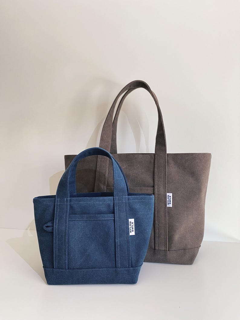 May include: A small blue denim tote bag and a larger brown canvas tote bag, both featuring two top handles, a front pocket, and a white rectangular label with the black text NYLON SMILE stitched onto the lower right side of each bag.