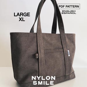 Könnte beinhalten: Eine große XL-Tragetasche in Dunkelbraun mit zwei Schulterriemen, einer Vordertasche und einem kleinen seitlichen Etikett. Der Text auf dem Bild lautet: PDF PATTERN, ENGLISH, ESPAÑOL, LARGE XL und NYLON SMILE. Die Tasche hat eine Stoffstruktur.