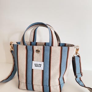 May include: A small tote bag featuring vertical stripes in light blue, dark brown, and off-white. The bag has two top handles, a detachable shoulder strap with gold-tone hardware, a central snap button closure, and a white label reading NYLON SMILE.