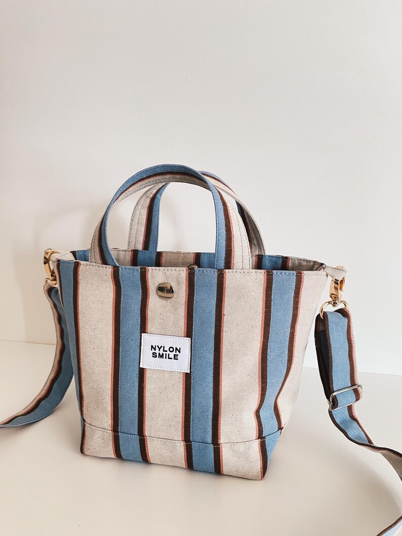 May include: A small tote bag featuring vertical stripes in light blue, dark brown, and off-white. The bag has two top handles, a detachable shoulder strap with gold-tone hardware, a central snap button closure, and a white label reading NYLON SMILE.