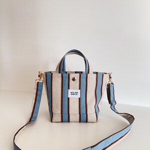 May include: A small tote bag featuring vertical stripes in light blue, dark brown, and beige. The bag has two top handles, a detachable shoulder strap, and a white rectangular label on the front with the black text NYLON SMILE.
