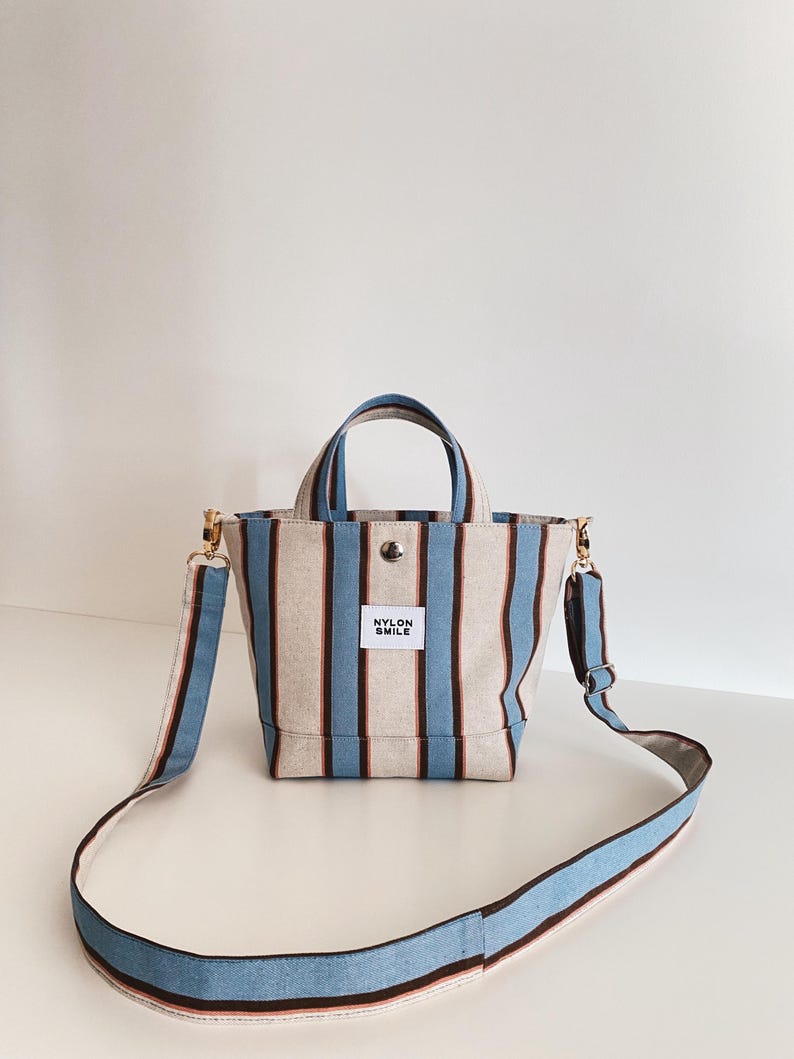 May include: A small tote bag featuring vertical stripes in light blue, dark brown, and beige. The bag has two top handles, a detachable shoulder strap, and a white rectangular label on the front with the black text NYLON SMILE.