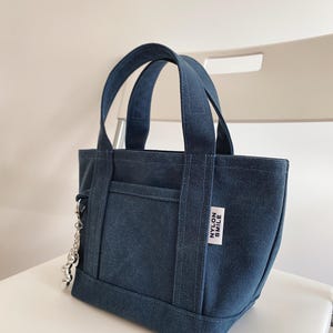 K&ouml;nnte beinhalten: Eine dunkelblaue Canvas-Tragetasche mit zwei stabilen Henkeln und einer Fronttasche. Ein wei&szlig;es, rechteckiges Stoffetikett mit dem schwarzen Text NYLON SMILE ist an der Seite angen&auml;ht. Ein silberfarbener Metallkettenanh&auml;nger h&auml;ngt an der Seite.
