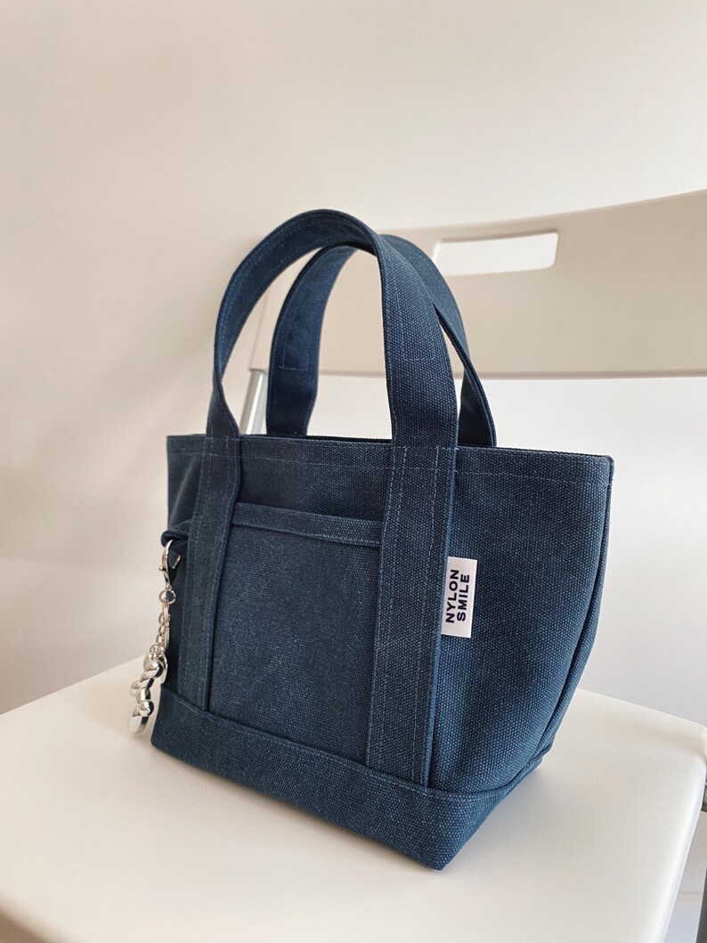 K&ouml;nnte beinhalten: Eine dunkelblaue Canvas-Tragetasche mit zwei stabilen Henkeln und einer Fronttasche. Ein wei&szlig;es, rechteckiges Stoffetikett mit dem schwarzen Text NYLON SMILE ist an der Seite angen&auml;ht. Ein silberfarbener Metallkettenanh&auml;nger h&auml;ngt an der Seite.