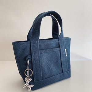 K&ouml;nnte beinhalten: Eine dunkelblaue Tragetasche aus Denim mit zwei stabilen Henkeln und einer Au&szlig;entasche. Ein silberfarbener Schl&uuml;sselanh&auml;nger in Form eines Ballonhundes ist an der Seite befestigt. Ein kleines wei&szlig;es Etikett mit dem Wort SMILE ist seitlich sichtbar.
