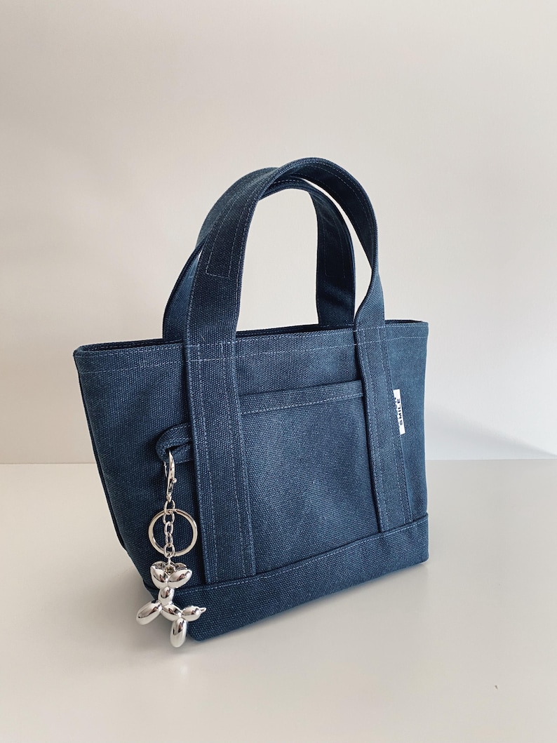 K&ouml;nnte beinhalten: Eine dunkelblaue Tragetasche aus Denim mit zwei stabilen Henkeln und einer Au&szlig;entasche. Ein silberfarbener Schl&uuml;sselanh&auml;nger in Form eines Ballonhundes ist an der Seite befestigt. Ein kleines wei&szlig;es Etikett mit dem Wort SMILE ist seitlich sichtbar.