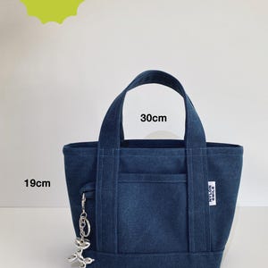 May include: A dark blue canvas tote bag with two top handles, a front pocket, and a silver balloon dog keychain. The bag features a white label with the text NYLON SMILE. Text indicates a skill level of beginner and dimensions of 30cm by 19cm.