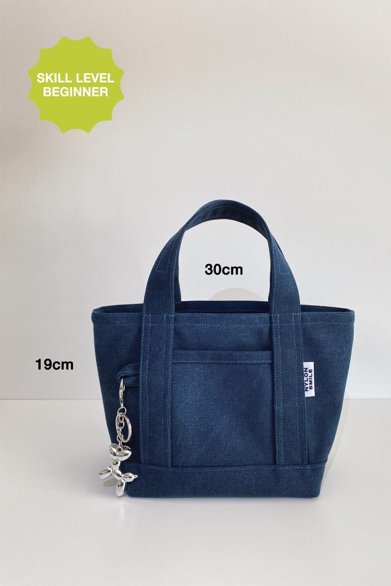 May include: A dark blue canvas tote bag with two top handles, a front pocket, and a silver balloon dog keychain. The bag features a white label with the text NYLON SMILE. Text indicates a skill level of beginner and dimensions of 30cm by 19cm.