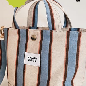 May include: A tote bag featuring vertical stripes in light blue, dark brown, and cream. The bag has two top handles, a silver snap button closure, and a white rectangular label with the black text NYLON SMILE. Text in the image reads INSTANT DOWNLOAD, LETTER, A4, A0.