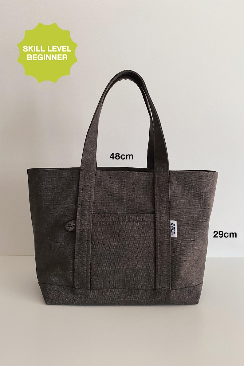 May include: A dark gray canvas tote bag with two shoulder straps, a front pocket, and a small white label reading NYLON SMILE. The bag measures 48 cm in width and 29 cm in height. A lime green starburst graphic in the top left corner reads SKILL LEVEL BEGINNER.