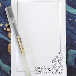 Op de afbeelding: Een wit notitieboekje met een zwarte rand en een zwarte pen. Het notitieboekje heeft een tekening van dobbelstenen in de rechterbenedenhoek.