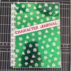 Puede incluir: Un diario de personaje verde con una encuadernación en espiral blanca y una etiqueta roja y blanca que dice "CHARACTER JOURNAL". La portada está decorada con un patrón repetido de dados blancos.
