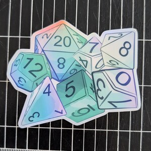 May include: A sticker featuring a collection of polyhedral dice in various shapes. The dice are primarily teal and blue with iridescent highlights, and each die displays its numerical values. The sticker is designed for tabletop gaming enthusiasts.