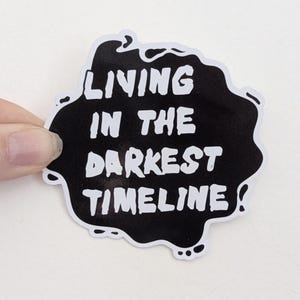 May include: Black and white sticker with the text "Living in the darkest timeline".