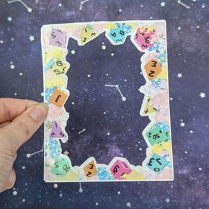 May include: A colorful sticker with a clear center, decorated with dice illustrations. The dice are various colors and feature numbers. The sticker has a textured, glittery border in pastel colors. The background is a starry night sky.