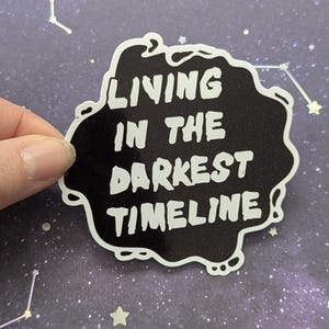 May include: Black and white sticker with the text "Living in the darkest timeline" on a black background with a white outline. The sticker is on a purple background with white glitter stars.