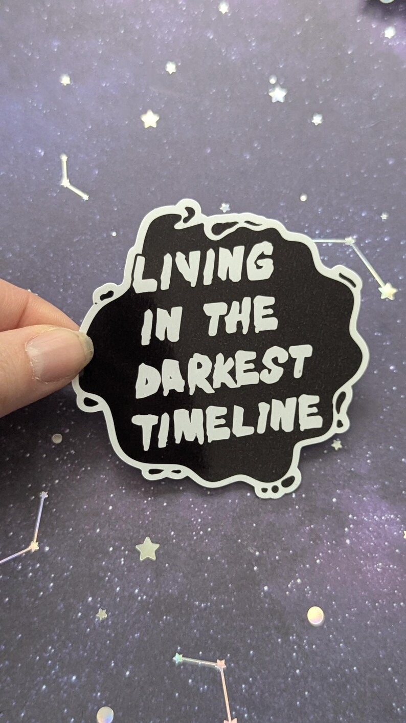 May include: Black and white sticker with the text "Living in the darkest timeline" on a black background with a white outline. The sticker is on a purple background with white glitter stars.