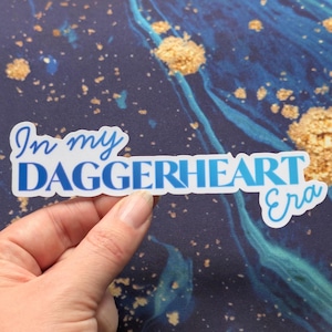 Pode incluir: Um autocolante branco com texto azul que diz "In my DAGGERHEART Era". O autocolante tem um efeito de gradiente azul. O fundo é azul escuro com manchas e redemoinhos dourados.