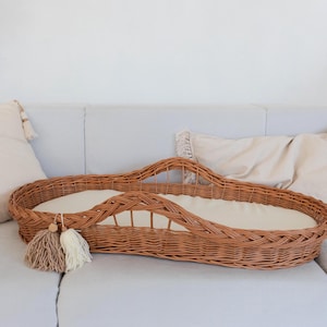 May include: A woven wicker bassinet with a white cotton liner. The bassinet is sitting on a white couch with pillows.
