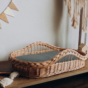Peut inclure: Un berceau en osier tressé avec une doublure en tissu gris. Le berceau est de forme ovale et possède un bord surélevé. Il est posé sur une surface en bois.