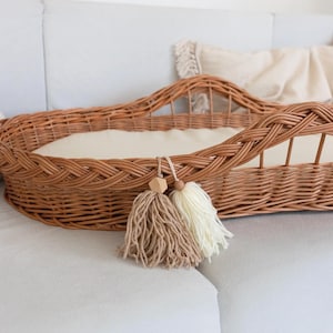 May include: A woven wicker bassinet in a natural brown color, featuring a braided edge and a soft, cream-colored interior. Two tassels, one beige and one cream, hang from the front. The bassinet is placed on a light gray surface.