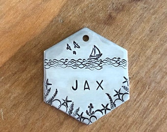 May include: A hexagonal silver-toned metal pet tag featuring a stamped nautical design. The top section shows a sailboat and birds over waves. The center is stamped with the name JAX. The bottom border displays starfish, seaweed, and an anchor.