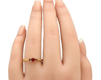 Puede incluir: Un anillo de oro con tres piedras preciosas rojas. El anillo tiene una banda de cuentas y una piedra central ovalada flanqueada por dos piedras redondas más pequeñas. El anillo se muestra en una mano con uñas cuidadas.