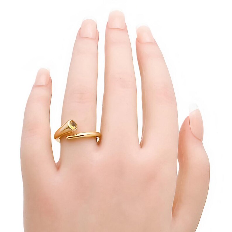 May include: A gold-colored ring with a spiral design and a yellow gemstone. The ring is worn on a finger, showcasing its elegant and modern style. The gemstone is round and set in a prong setting, adding a touch of sparkle.