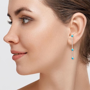 May include: A silver drop earring featuring a thin vertical metal bar accented with three small, round, bright turquoise stones spaced evenly along the length of the post. The earring hangs straight down from the earlobe in a linear design.